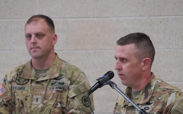 Two U.S. Army soldiers in OCP (Operational Camouflage Pattern) uniforms are participating in a formal military ceremony or briefing. One soldier is speaking at a podium with a microphone, while the other stands attentively in the background. Name tapes visible on their uniforms read 'DAUMER' and partially visible on the speaker.