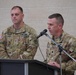 Two U.S. Army soldiers in OCP (Operational Camouflage Pattern) uniforms are participating in a formal military ceremony or briefing. One soldier is speaking at a podium with a microphone, while the other stands attentively in the background. Name tapes visible on their uniforms read 'DAUMER' and partially visible on the speaker.