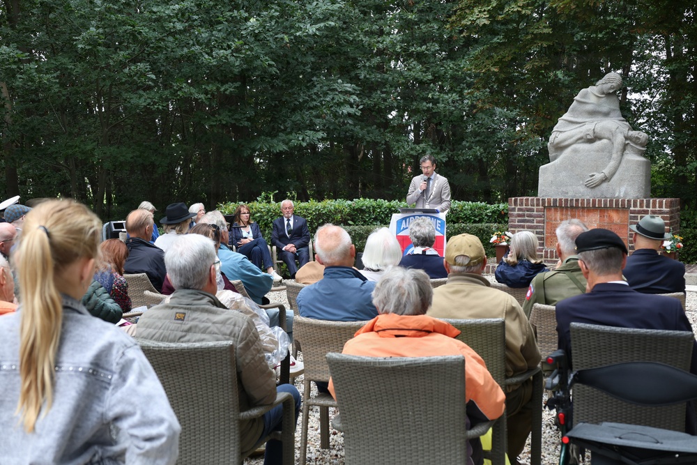 An outdoor memorial ceremony is taking place in what appears to be a garden or park setting. A speaker addresses a seated audience from a podium bearing an 'AIRBORNE' sign, with a stone memorial statue visible in the background. The gathering appears to be a commemorative event, likely related to WWII Airborne operations, attended by a mixed-age crowd including some individuals in military attire.