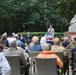 An outdoor memorial ceremony is taking place in what appears to be a garden or park setting. A speaker addresses a seated audience from a podium bearing an 'AIRBORNE' sign, with a stone memorial statue visible in the background. The gathering appears to be a commemorative event, likely related to WWII Airborne operations, attended by a mixed-age crowd including some individuals in military attire.