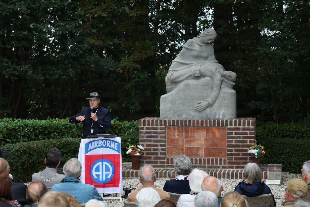 An outdoor memorial ceremony is taking place at what appears to be a war memorial or monument. A speaker stands at a podium displaying the 82nd Airborne Division insignia, addressing a seated audience of mostly elderly attendees. The setting includes a large stone sculpture monument on a brick base, surrounded by hedges and trees.