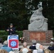 An outdoor memorial ceremony is taking place at what appears to be a war memorial or monument. A speaker stands at a podium displaying the 82nd Airborne Division insignia, addressing a seated audience of mostly elderly attendees. The setting includes a large stone sculpture monument on a brick base, surrounded by hedges and trees.