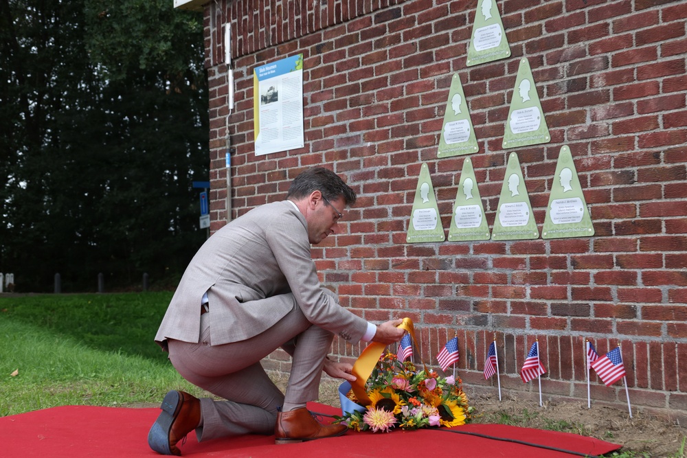 A man in a light gray suit is kneeling on a red carpet to place a floral wreath at the base of a brick memorial wall. The wall features multiple triangular green plaques arranged in a pyramid pattern, each bearing silhouette profiles and inscriptions. Small American flags are planted along the base of the wall, indicating a commemorative or dedication ceremony.