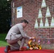 A man in a light gray suit is kneeling on a red carpet to place a floral wreath at the base of a brick memorial wall. The wall features multiple triangular green plaques arranged in a pyramid pattern, each bearing silhouette profiles and inscriptions. Small American flags are planted along the base of the wall, indicating a commemorative or dedication ceremony.