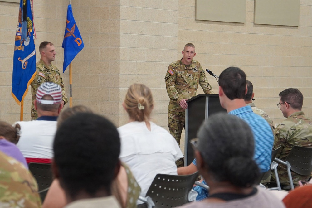 A military officer in Army Combat Uniform (ACU/OCP pattern) is delivering a speech at a podium during what appears to be an official military ceremony or briefing. A second uniformed soldier stands at attention near two flags in the background. An audience of both military personnel and civilians is seated facing the speaker.