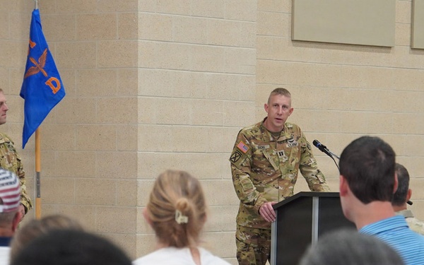 A military officer in Army Combat Uniform (ACU/OCP pattern) is delivering a speech at a podium during what appears to be an official military ceremony or briefing. A second uniformed soldier stands at attention near two flags in the background. An audience of both military personnel and civilians is seated facing the speaker.