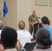 A military officer in Army Combat Uniform (ACU/OCP pattern) is delivering a speech at a podium during what appears to be an official military ceremony or briefing. A second uniformed soldier stands at attention near two flags in the background. An audience of both military personnel and civilians is seated facing the speaker.