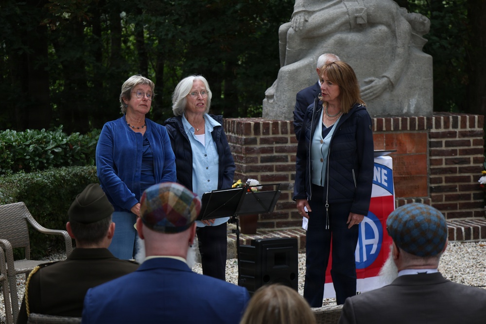 Three women stand at a podium during what appears to be a formal outdoor memorial or commemorative ceremony. Behind them is a stone monument on a brick pedestal, alongside a banner featuring what appears to be the 82nd Airborne Division insignia. Audience members, including individuals wearing military caps and kippot, are seated in the foreground.
