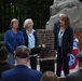 Three women stand at a podium during what appears to be a formal outdoor memorial or commemorative ceremony. Behind them is a stone monument on a brick pedestal, alongside a banner featuring what appears to be the 82nd Airborne Division insignia. Audience members, including individuals wearing military caps and kippot, are seated in the foreground.