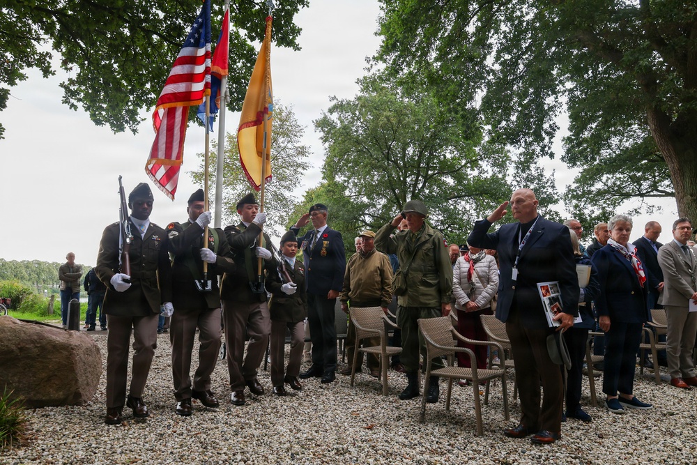 A military memorial or commemorative ceremony is taking place outdoors, featuring a color guard carrying multiple flags including the American flag. Attendees in both military uniforms and civilian dress are saluting, while a crowd of observers stands in the background. The setting appears to be a park or memorial ground with gravel surface and large trees.