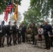 A military memorial or commemorative ceremony is taking place outdoors, featuring a color guard carrying multiple flags including the American flag. Attendees in both military uniforms and civilian dress are saluting, while a crowd of observers stands in the background. The setting appears to be a park or memorial ground with gravel surface and large trees.