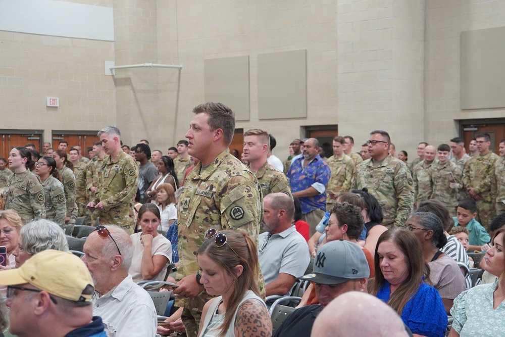 A military ceremony or formation is taking place indoors, likely at a U.S. Army installation. Soldiers in OCP (Operational Camouflage Pattern) uniforms are standing at attention or in formation while civilian family members and guests are seated in chairs in the foreground. The setting appears to be a large assembly hall or auditorium on a military base.