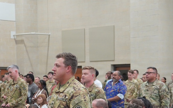 A military ceremony or formation is taking place indoors, likely at a U.S. Army installation. Soldiers in OCP (Operational Camouflage Pattern) uniforms are standing at attention or in formation while civilian family members and guests are seated in chairs in the foreground. The setting appears to be a large assembly hall or auditorium on a military base.