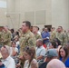 A military ceremony or formation is taking place indoors, likely at a U.S. Army installation. Soldiers in OCP (Operational Camouflage Pattern) uniforms are standing at attention or in formation while civilian family members and guests are seated in chairs in the foreground. The setting appears to be a large assembly hall or auditorium on a military base.