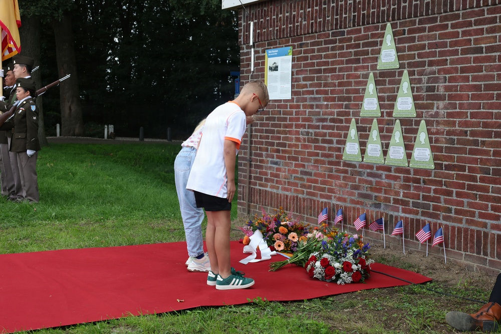 A memorial ceremony is taking place at a brick wall monument featuring triangular green plaques with silhouettes. Two children are bowing respectfully before floral tributes and small American flags placed on a red carpet. A military honor guard with rifles and a flag standard is visible in the background.