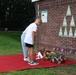 A memorial ceremony is taking place at a brick wall monument featuring triangular green plaques with silhouettes. Two children are bowing respectfully before floral tributes and small American flags placed on a red carpet. A military honor guard with rifles and a flag standard is visible in the background.