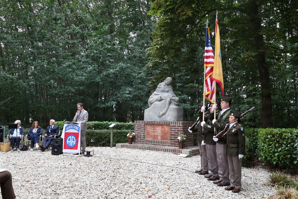 A formal military memorial ceremony is taking place outdoors at a monument associated with the 82nd Airborne Division ('All American'). A speaker addresses attendees from a podium bearing the 82nd Airborne patch, while a color guard stands at attention holding the American flag and what appears to be a unit or state flag. A stone sculpture of a mourning figure sits atop a brick pedestal in the background.