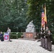 A formal military memorial ceremony is taking place outdoors at a monument associated with the 82nd Airborne Division ('All American'). A speaker addresses attendees from a podium bearing the 82nd Airborne patch, while a color guard stands at attention holding the American flag and what appears to be a unit or state flag. A stone sculpture of a mourning figure sits atop a brick pedestal in the background.