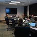 Military personnel in camouflage uniforms are gathered in a conference room setting, appearing to conduct a briefing or operational planning session. Multiple individuals are seated around a large conference table equipped with laptops, phones, and documents, while one person stands and appears to be addressing the group. A large display screen and a digital clock/counter are visible on the wall behind them.