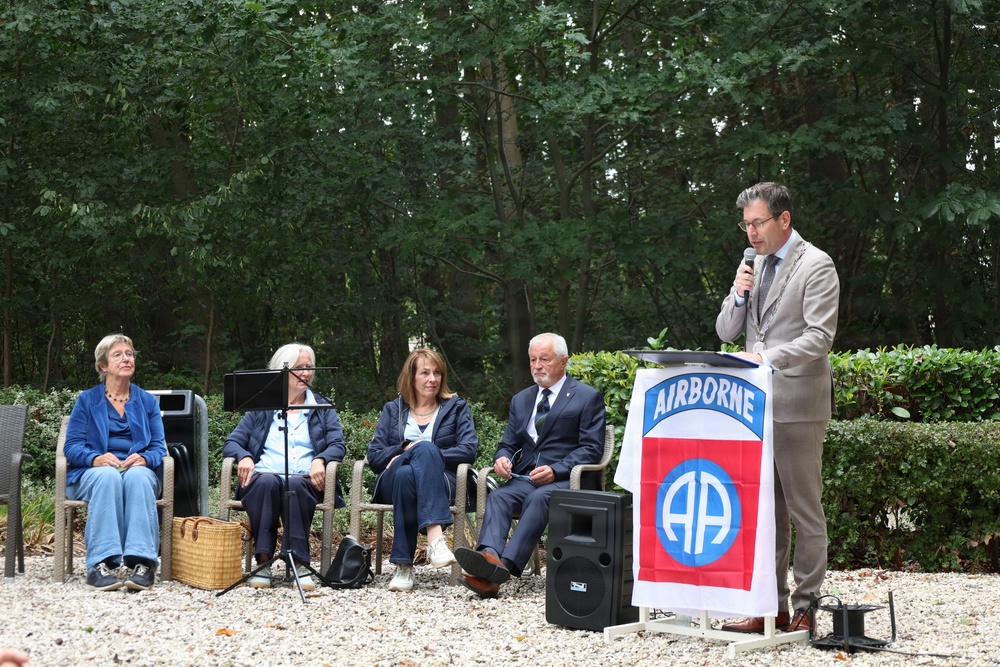 An outdoor ceremony or commemorative event featuring a speaker at a podium adorned with the 82nd Airborne Division flag. Four individuals are seated in chairs to the left, while a man in a suit speaks into a microphone at the podium. The setting appears to be a garden or park with gravel ground and dense green trees in the background.