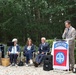 An outdoor ceremony or commemorative event featuring a speaker at a podium adorned with the 82nd Airborne Division flag. Four individuals are seated in chairs to the left, while a man in a suit speaks into a microphone at the podium. The setting appears to be a garden or park with gravel ground and dense green trees in the background.
