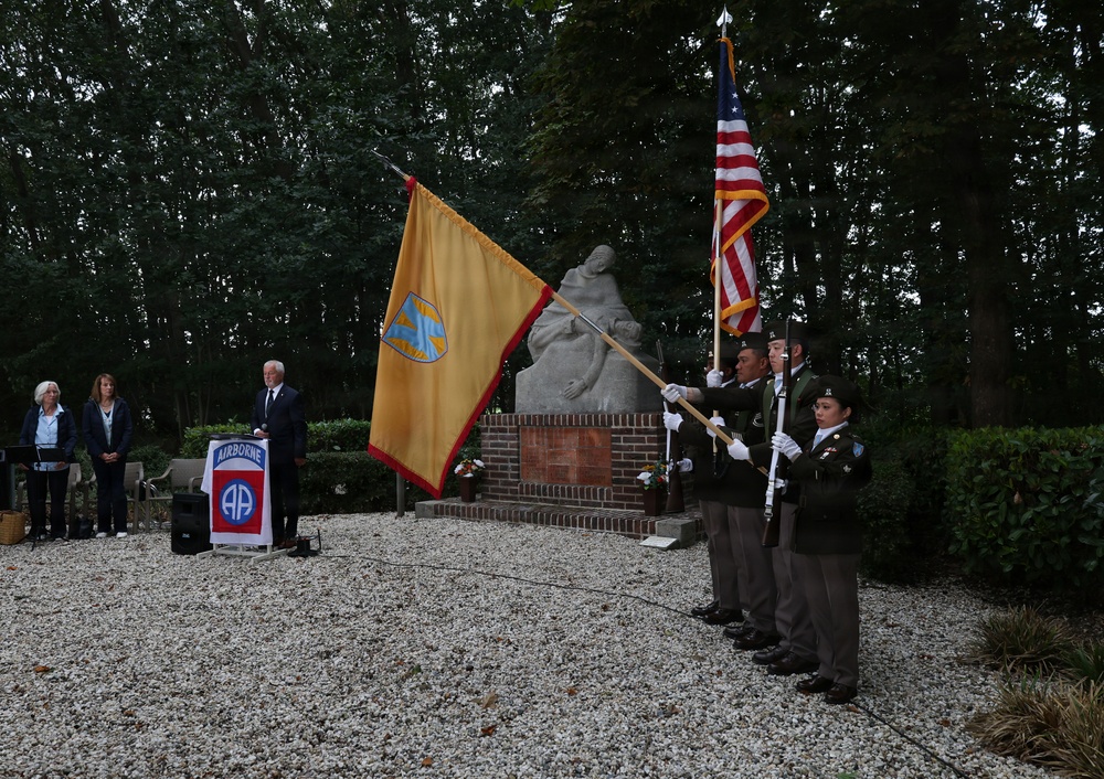 A formal military memorial ceremony is taking place outdoors at a monument, with a color guard presenting flags including the American flag and a gold unit guidon. A speaker stands at a podium bearing the 82nd Airborne Division patch, while attendees observe the ceremony. The setting appears to be a commemorative site, likely in the Netherlands given the architectural and landscape style.
