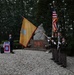 A formal military memorial ceremony is taking place outdoors at a monument, with a color guard presenting flags including the American flag and a gold unit guidon. A speaker stands at a podium bearing the 82nd Airborne Division patch, while attendees observe the ceremony. The setting appears to be a commemorative site, likely in the Netherlands given the architectural and landscape style.