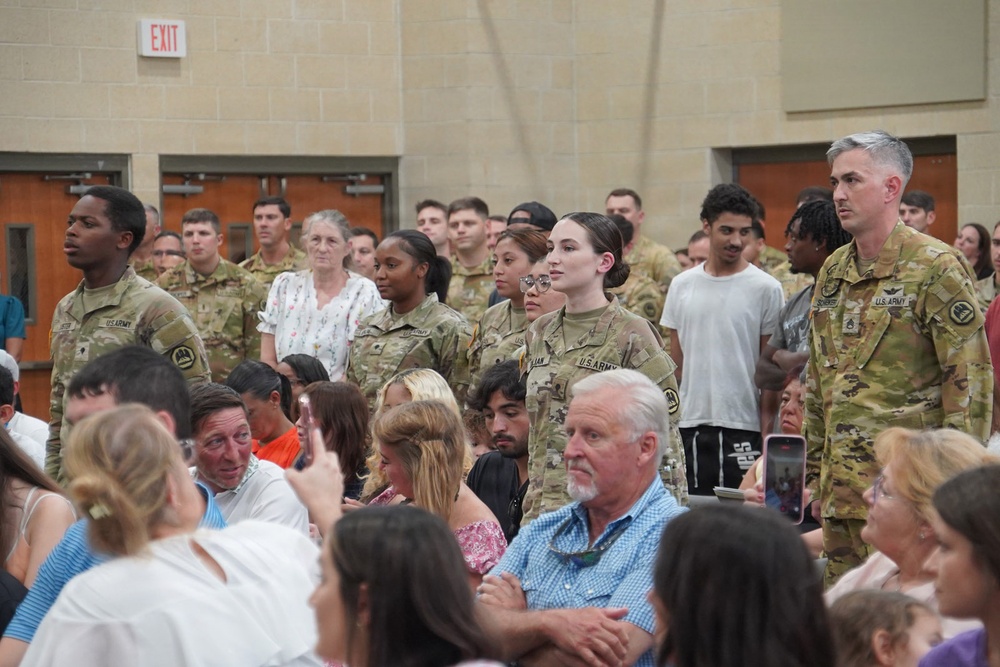 A group of U.S. Army soldiers in OCP (Operational Camouflage Pattern) uniforms are standing at attention or in formation during what appears to be a military ceremony or event. They are surrounded by seated civilian attendees, likely family members and guests, in what looks like an indoor venue or gymnasium. The event appears to be a formal military occasion such as a graduation, promotion ceremony, or unit activation.