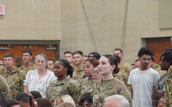 A group of U.S. Army soldiers in OCP (Operational Camouflage Pattern) uniforms are standing at attention or in formation during what appears to be a military ceremony or event. They are surrounded by seated civilian attendees, likely family members and guests, in what looks like an indoor venue or gymnasium. The event appears to be a formal military occasion such as a graduation, promotion ceremony, or unit activation.