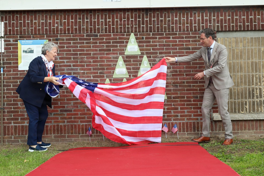 Two individuals are unveiling a memorial or commemorative installation on a brick wall by removing a large American flag used as a covering. The ceremony takes place on a red carpet in front of a brick building, with several triangular green plaques mounted on the wall and small American flags placed along the ground. The setting and attire suggest an official commemorative event, likely related to American military history or transatlantic relations.