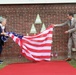 Two individuals are unveiling a memorial or commemorative installation on a brick wall by removing a large American flag used as a covering. The ceremony takes place on a red carpet in front of a brick building, with several triangular green plaques mounted on the wall and small American flags placed along the ground. The setting and attire suggest an official commemorative event, likely related to American military history or transatlantic relations.