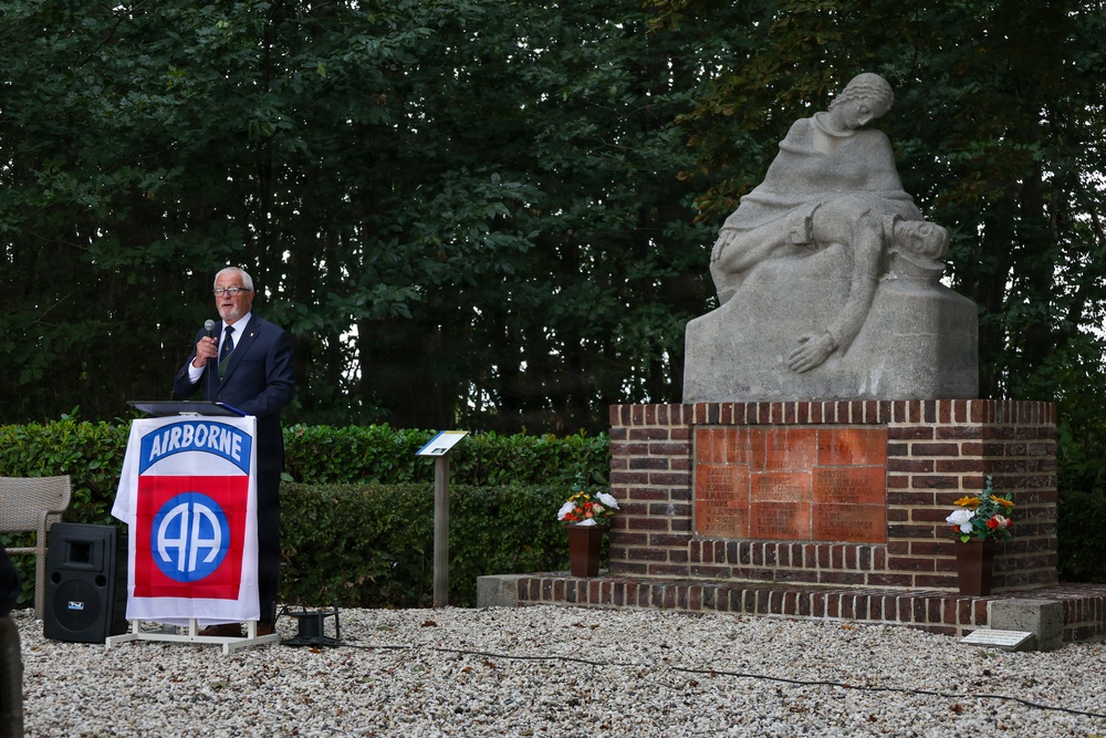 A man in a dark suit is speaking at a podium draped with an 82nd Airborne Division flag during what appears to be a memorial ceremony. Behind him stands a stone war memorial sculpture depicting a mourning figure holding a fallen soldier, mounted on a brick pedestal with inscribed plaques. Floral tributes are placed at the base of the monument.