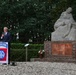 A man in a dark suit is speaking at a podium draped with an 82nd Airborne Division flag during what appears to be a memorial ceremony. Behind him stands a stone war memorial sculpture depicting a mourning figure holding a fallen soldier, mounted on a brick pedestal with inscribed plaques. Floral tributes are placed at the base of the monument.