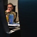 A military personnel member in camouflage uniform is seated at a desk, viewed through a gap between computer monitors. The individual appears to be in a thoughtful or attentive posture with hand raised to chin. The setting appears to be a military office or operations center environment.