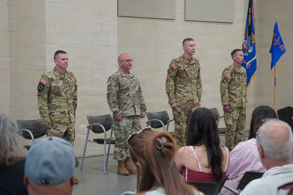 Four U.S. Army soldiers in OCP (Operational Camouflage Pattern) uniforms stand at attention during what appears to be a military ceremony, likely a change of command or awards ceremony. They are positioned in front of a seated audience of civilians, with military guidon flags visible in the background. The setting appears to be an indoor military facility or armory.