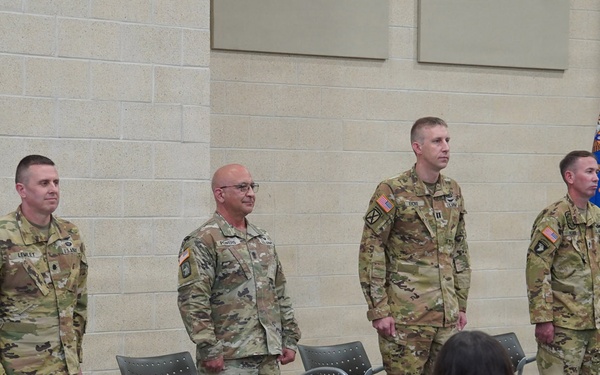 Four U.S. Army soldiers in OCP (Operational Camouflage Pattern) uniforms stand at attention during what appears to be a military ceremony, likely a change of command or awards ceremony. They are positioned in front of a seated audience of civilians, with military guidon flags visible in the background. The setting appears to be an indoor military facility or armory.