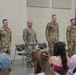 Four U.S. Army soldiers in OCP (Operational Camouflage Pattern) uniforms stand at attention during what appears to be a military ceremony, likely a change of command or awards ceremony. They are positioned in front of a seated audience of civilians, with military guidon flags visible in the background. The setting appears to be an indoor military facility or armory.