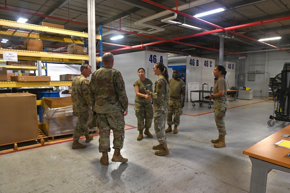 A group of military personnel in OCP (Operational Camouflage Pattern) uniforms are gathered inside a large warehouse or logistics facility. They appear to be conducting a briefing, inspection, or tour of the storage area. The facility contains numbered storage bays (46A, 46B, 46C, 46D) and shelving units with boxes and palletized cargo.