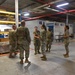 A group of military personnel in OCP (Operational Camouflage Pattern) uniforms are gathered inside a large warehouse or logistics facility. They appear to be conducting a briefing, inspection, or tour of the storage area. The facility contains numbered storage bays (46A, 46B, 46C, 46D) and shelving units with boxes and palletized cargo.
