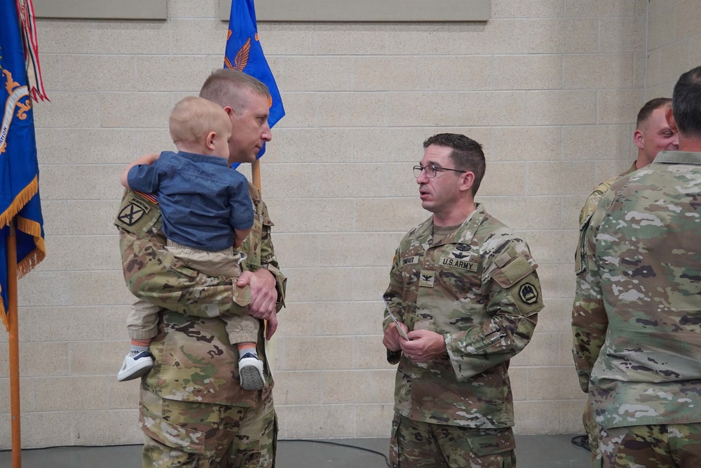 A military ceremony or informal gathering featuring U.S. Army soldiers in Operational Camouflage Pattern (OCP) uniforms. One soldier is holding a young infant/toddler while conversing with another soldier who wears colonel-rank insignia (eagle) and glasses. Military guidons and flags are visible in the background.