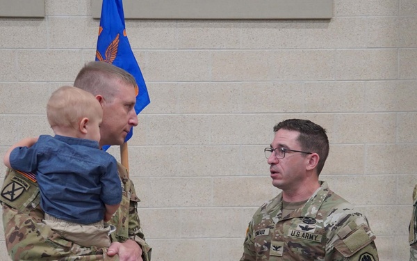 A military ceremony or informal gathering featuring U.S. Army soldiers in Operational Camouflage Pattern (OCP) uniforms. One soldier is holding a young infant/toddler while conversing with another soldier who wears colonel-rank insignia (eagle) and glasses. Military guidons and flags are visible in the background.