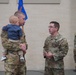 A military ceremony or informal gathering featuring U.S. Army soldiers in Operational Camouflage Pattern (OCP) uniforms. One soldier is holding a young infant/toddler while conversing with another soldier who wears colonel-rank insignia (eagle) and glasses. Military guidons and flags are visible in the background.