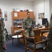 Three military personnel in OCP (Operational Camouflage Pattern) uniforms are gathered in what appears to be a military office. The central figure, standing near a desk, is holding documents and appears to be briefing or conversing with the other two individuals. The office contains personal photos, awards, and standard office furniture consistent with a senior military officer's workspace.