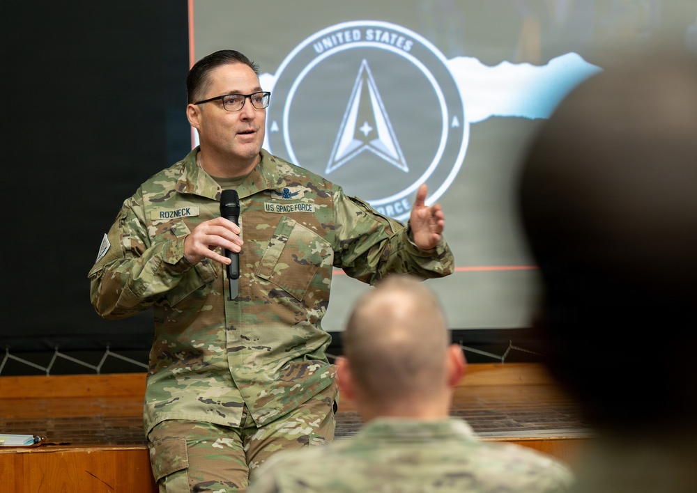 A U.S. Space Force member named Rozneck is delivering a presentation or briefing to an audience while holding a microphone. He is wearing OCP (Operational Camouflage Pattern) uniform with a 'U.S. SPACE FORCE' tape and a name tape reading 'ROZNECK'. Behind him is a projected slide displaying the United States Space Force seal/logo.