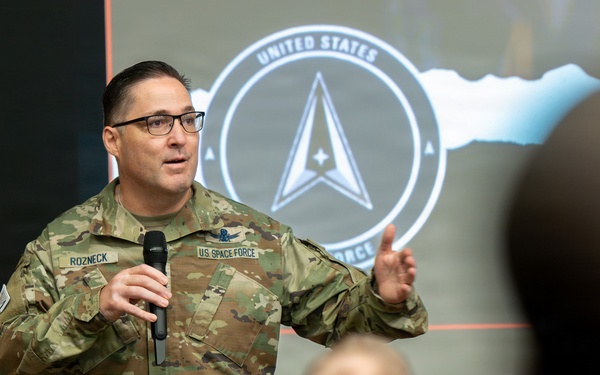 A U.S. Space Force member named Rozneck is delivering a presentation or briefing to an audience while holding a microphone. He is wearing OCP (Operational Camouflage Pattern) uniform with a 'U.S. SPACE FORCE' tape and a name tape reading 'ROZNECK'. Behind him is a projected slide displaying the United States Space Force seal/logo.