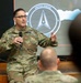 A U.S. Space Force member named Rozneck is delivering a presentation or briefing to an audience while holding a microphone. He is wearing OCP (Operational Camouflage Pattern) uniform with a 'U.S. SPACE FORCE' tape and a name tape reading 'ROZNECK'. Behind him is a projected slide displaying the United States Space Force seal/logo.