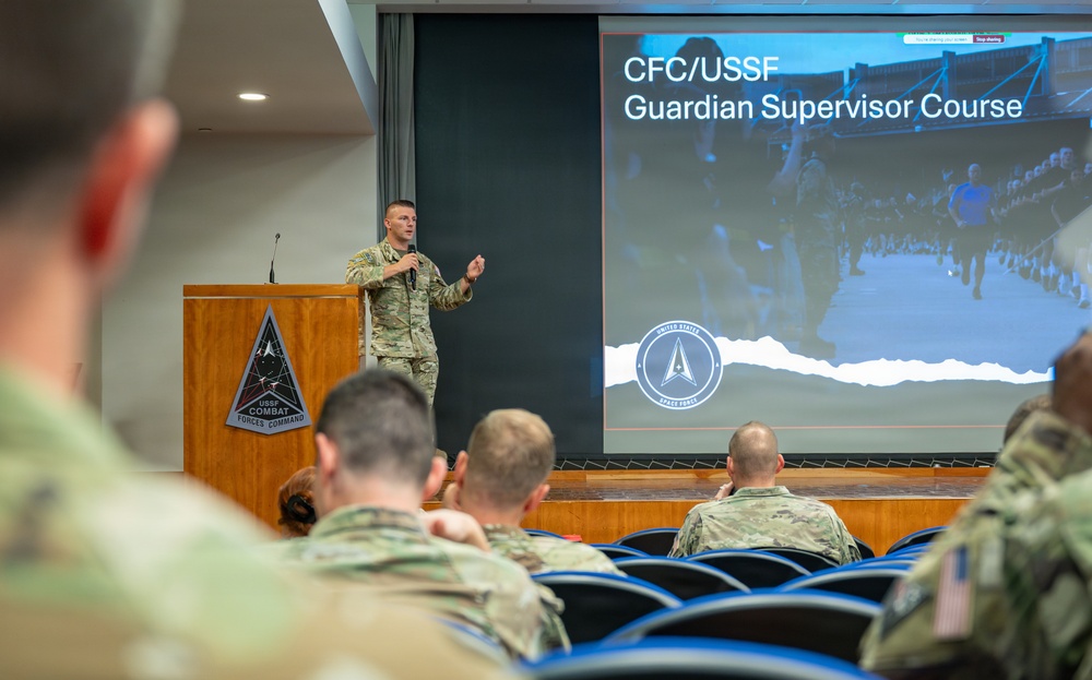 A military officer in camouflage uniform is presenting at a podium bearing the USSF Combat Forces Command emblem to an audience of uniformed personnel. The presentation slide behind him reads 'CFC/USSF Guardian Supervisor Course' and displays the United States Space Force logo. The setting appears to be a formal briefing room or auditorium.