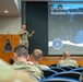 A military officer in camouflage uniform is presenting at a podium bearing the USSF Combat Forces Command emblem to an audience of uniformed personnel. The presentation slide behind him reads 'CFC/USSF Guardian Supervisor Course' and displays the United States Space Force logo. The setting appears to be a formal briefing room or auditorium.