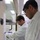 Two laboratory researchers are working inside or adjacent to a chemical fume hood in what appears to be a modern laboratory setting. Both individuals are wearing standard personal protective equipment including white lab coats and purple nitrile gloves, with one also wearing safety glasses. They appear to be collaboratively handling laboratory equipment or samples within the fume hood.