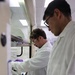 Two laboratory researchers are working inside or adjacent to a chemical fume hood in what appears to be a modern laboratory setting. Both individuals are wearing standard personal protective equipment including white lab coats and purple nitrile gloves, with one also wearing safety glasses. They appear to be collaboratively handling laboratory equipment or samples within the fume hood.